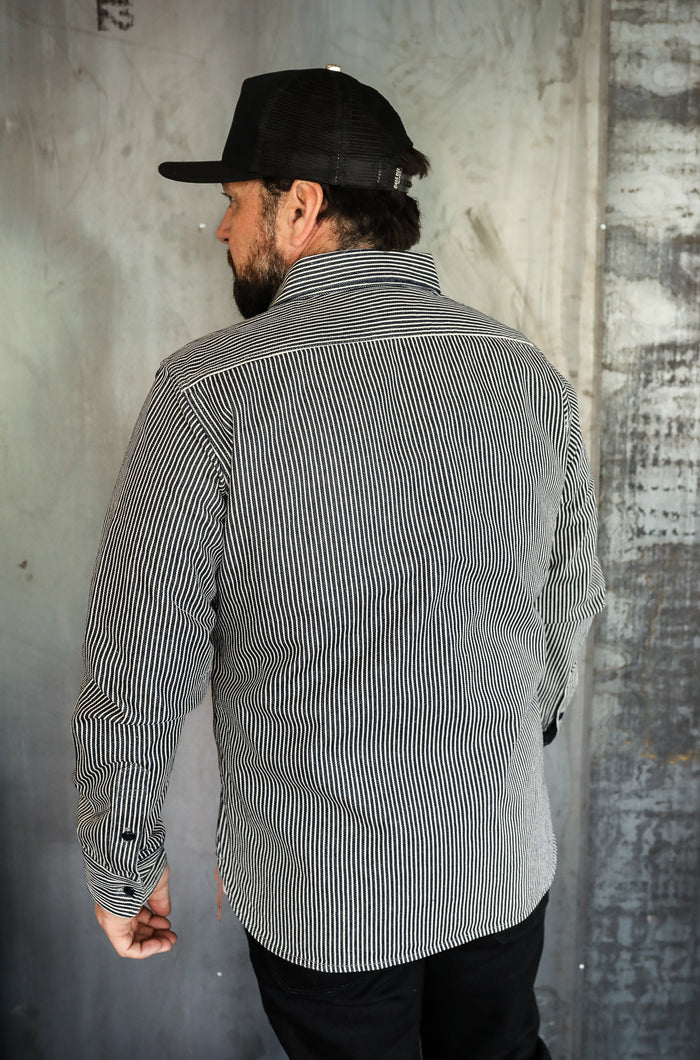 Man wearing black and white striped work shirt and black mesh cap standing in industrial setting