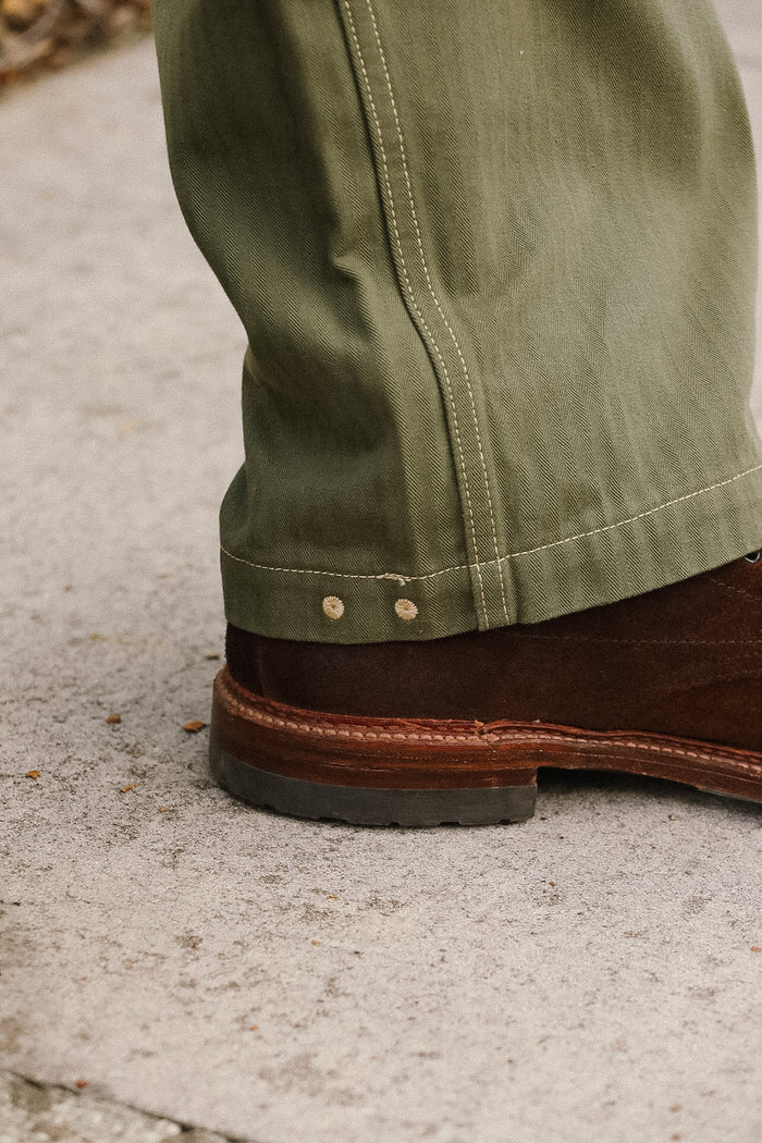 Close-up of green pants with a brown boot on a concrete surface