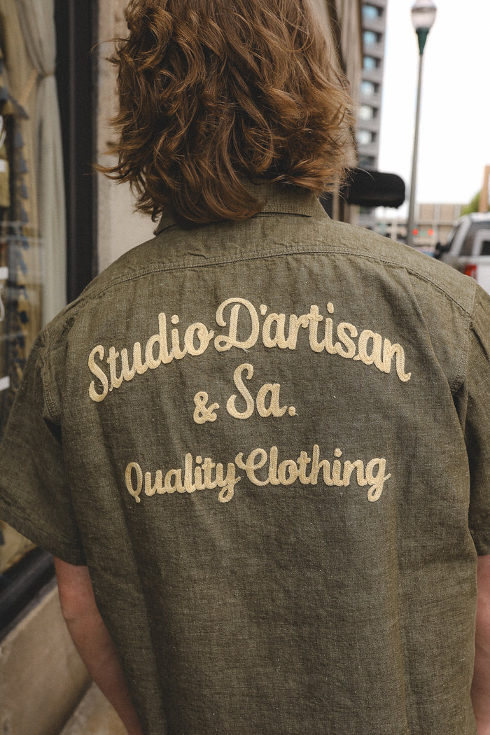 Back view of a person wearing an olive green shirt embroidered with 'Studio D'artisan & Sa. Quality Clothing' outdoors in a city street
