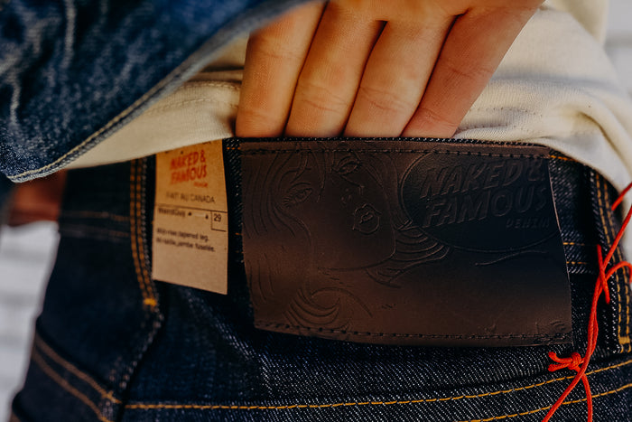 Close-up of hand showing Naked & Famous denim leather patch and fabric label on dark blue jeans