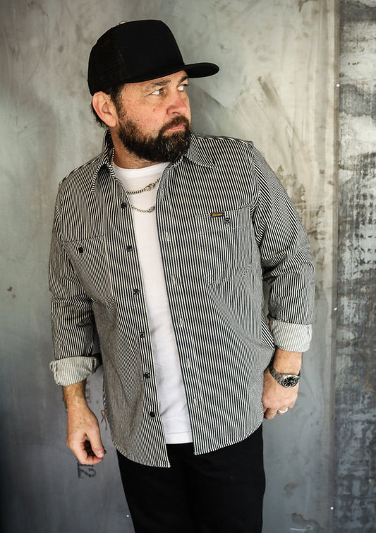 Man wearing black and white striped work shirt, white tee, black cap, and silver watch against industrial wall