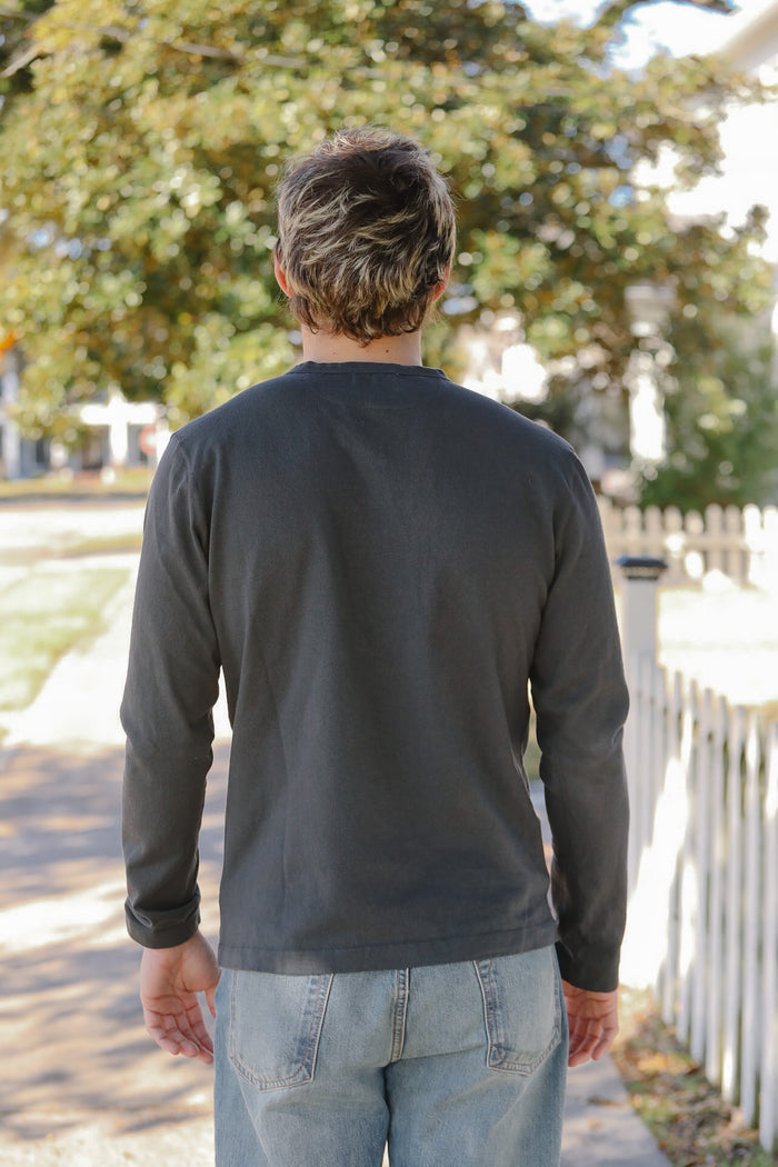 Man wearing a dark long-sleeve shirt and light blue jeans walking away from the camera with trees and a white fence in the background.