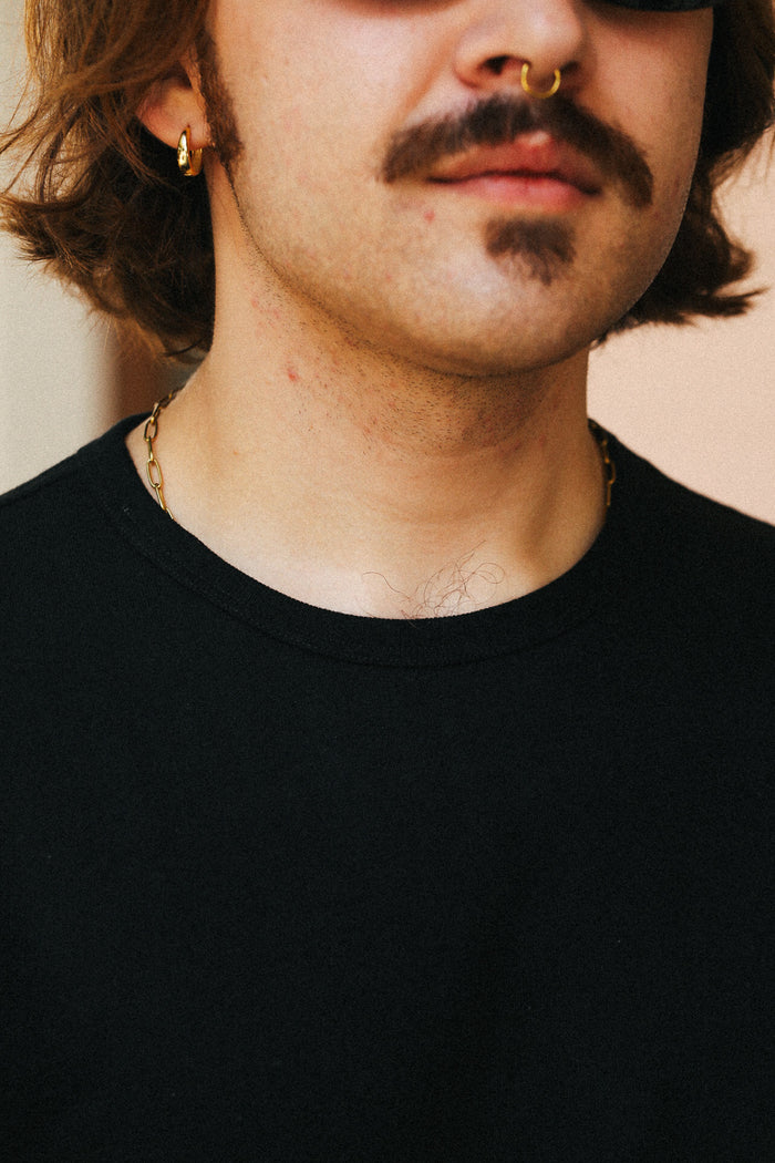 Close-up of a person wearing a black crew-neck t-shirt, a gold hoop earring, a nose ring, and a gothic mustache.