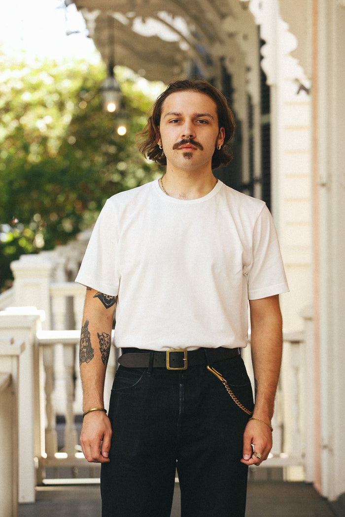 A man with a mustache and tattoos stands on a porch wearing a plain white crew-neck t-shirt, dark jeans, a black belt, and a gold chain accessory.