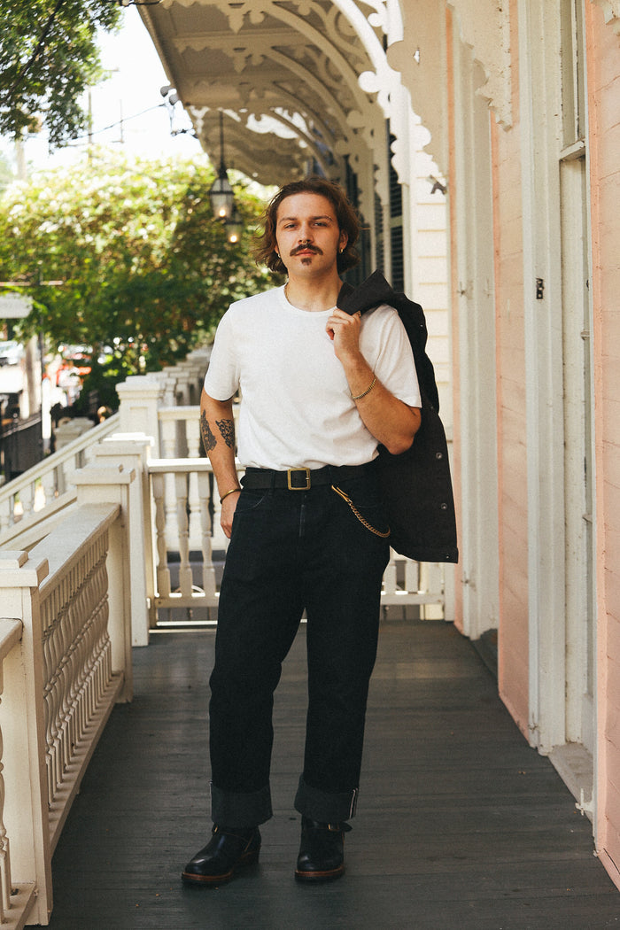 A man stands on a porch wearing a white crew-neck t-shirt, dark charcoal gray cuffed jeans, and black leather boots, with a black jacket slouched over his shoulder. The background features a white decorative porch railing and a pink building facade.
