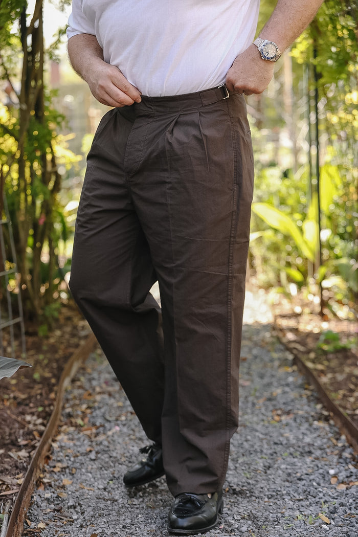 Person wearing brown pants and a white shirt standing on a gravel path with greenery in the background