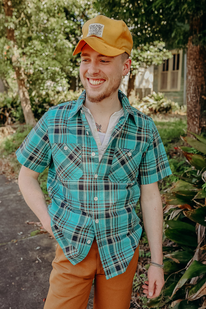 Smiling young man in a turquoise plaid short-sleeve shirt, mustard cap, and brown pants outdoors in a sunny garden setting