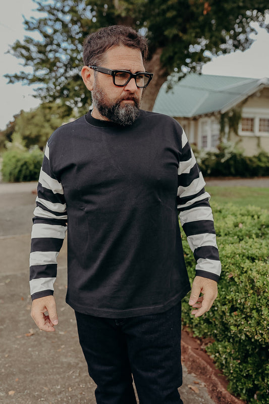 Man with beard and glasses wearing black shirt with black-and-white striped sleeves outdoors near a house