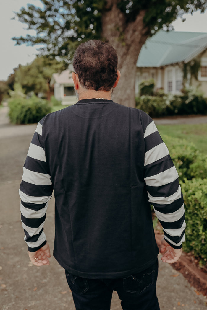 Man wearing black long-sleeve shirt with white stripes on sleeves, standing outdoors near bushes and a house