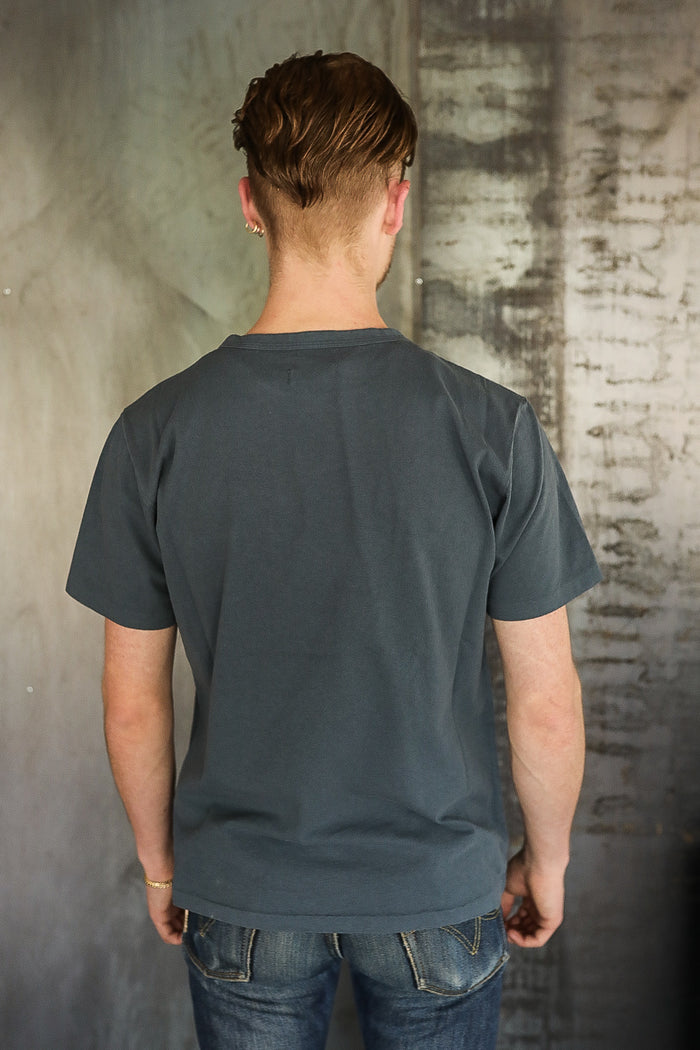 Man wearing a plain dark gray t-shirt and blue jeans standing with back to camera against textured wall