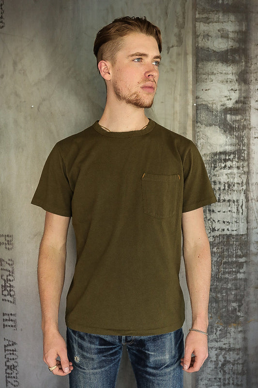 Man wearing olive green short sleeve pocket t-shirt and distressed blue jeans against textured concrete wall