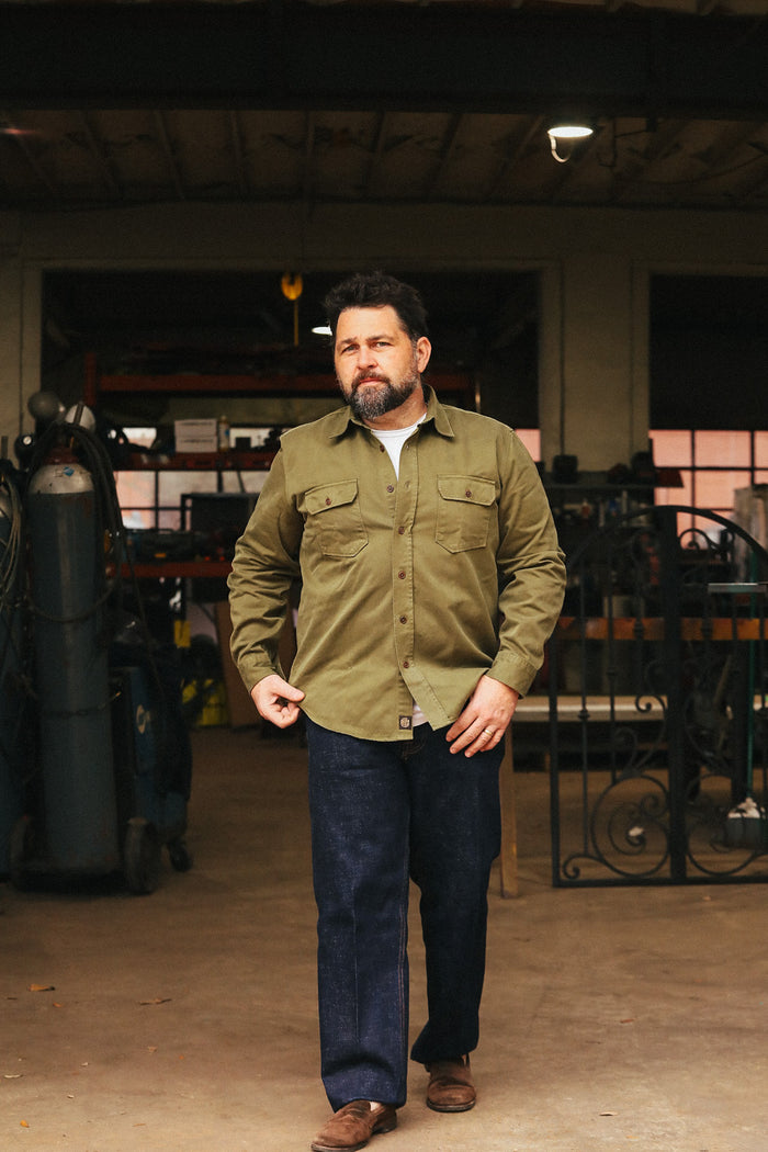 Man wearing a green jacket and blue jeans standing in an indoor setting with equipment in the background.