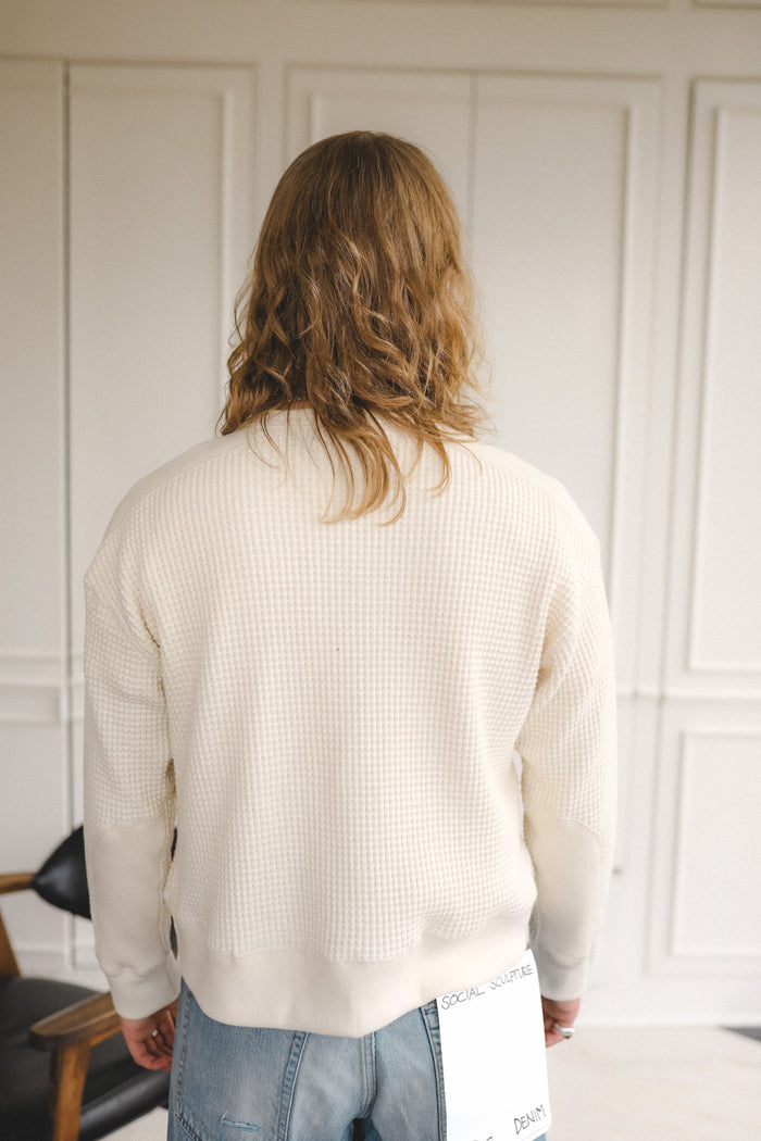 Person wearing a cream sweater and jeans standing in a room with white paneled walls.