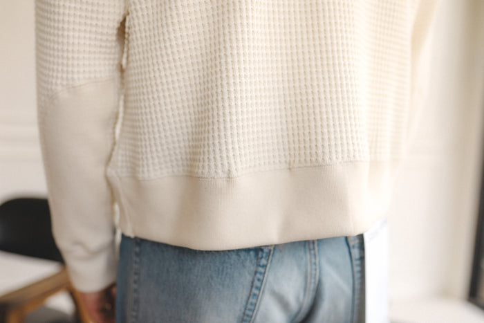 Person wearing a beige textured sweater and blue jeans indoors.