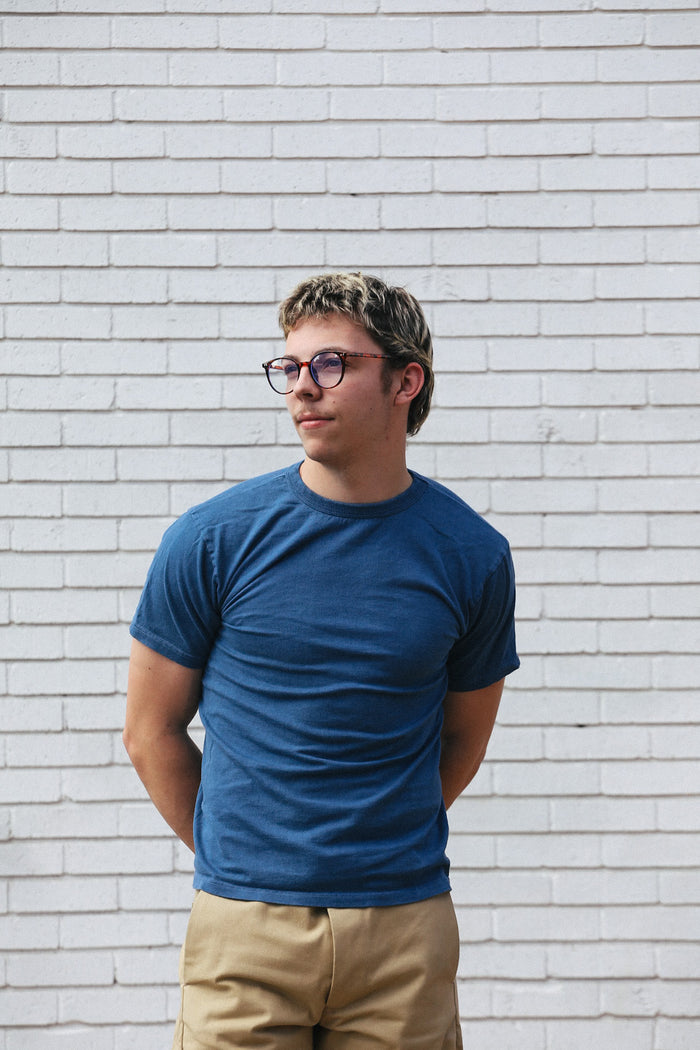 Young man with glasses wearing a blue t-shirt and beige pants standing against white brick wall