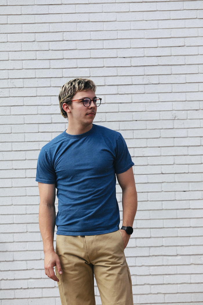 Young man wearing a blue t-shirt and beige pants standing against a white brick wall