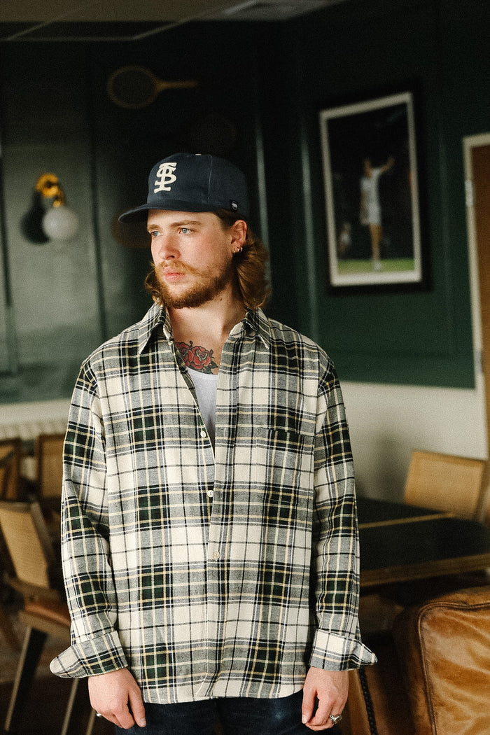 Man wearing a plaid shirt and cap in an indoor setting with framed pictures on the wall.