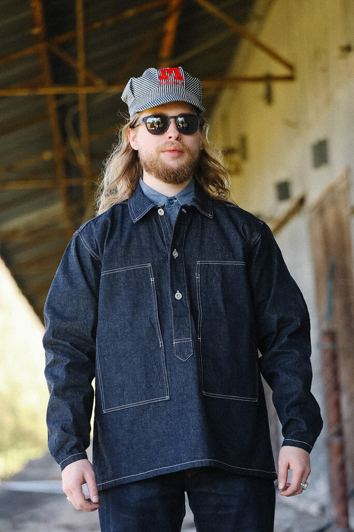 Man wearing a dark denim jacket and cap with a logo, standing in an industrial setting.