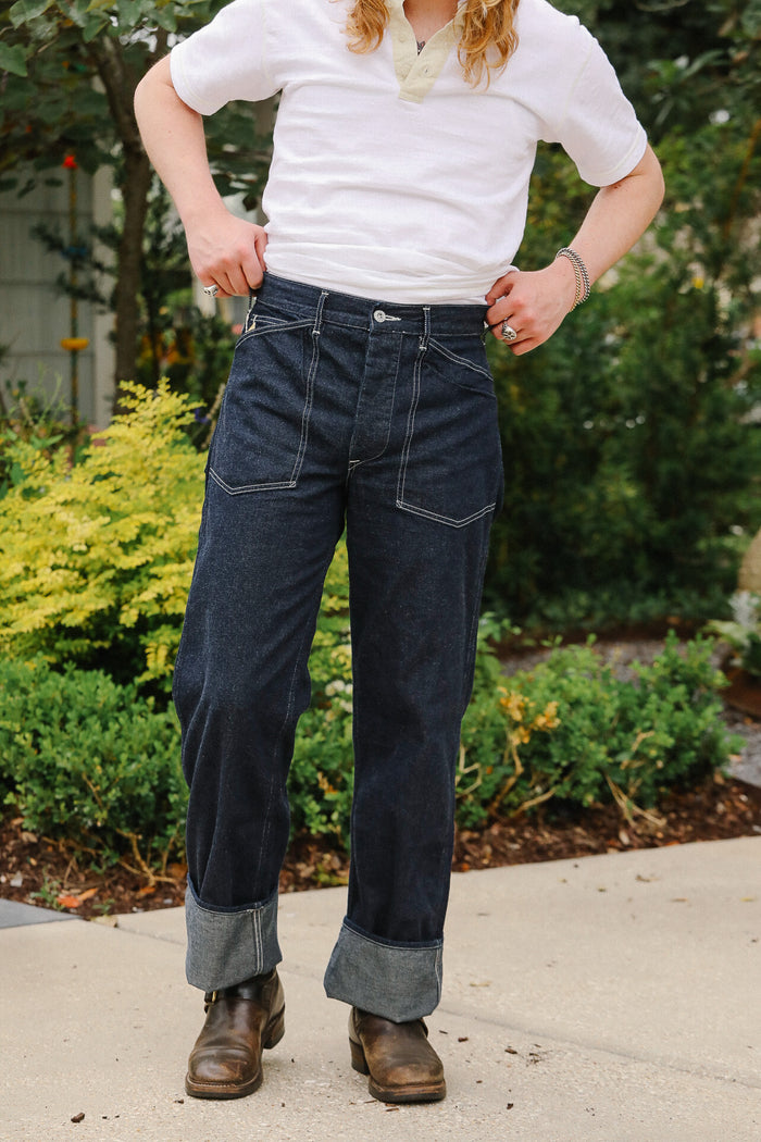 Person wearing dark blue jeans and a white shirt standing outdoors with greenery in the background