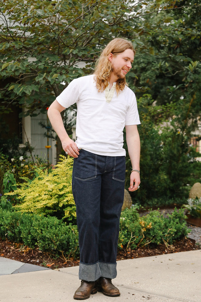 Person wearing a white t-shirt and dark jeans standing outdoors with greenery in the background