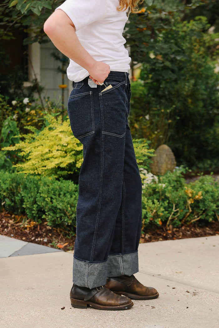 Person wearing dark blue jeans and brown shoes standing outdoors with greenery in the background