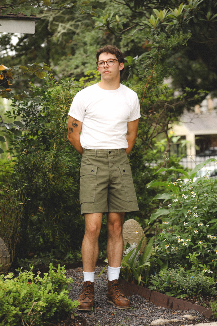 Man standing in a garden wearing a white t-shirt and green shorts.