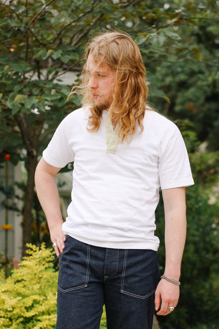 Person wearing a white t-shirt and dark jeans standing outdoors with greenery in the background