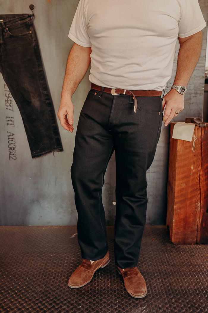 Man wearing dark denim jeans with brown belt and brown suede shoes in industrial setting