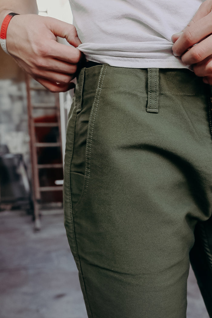 Close-up of person wearing olive green durable work pants and adjusting white shirt in workshop