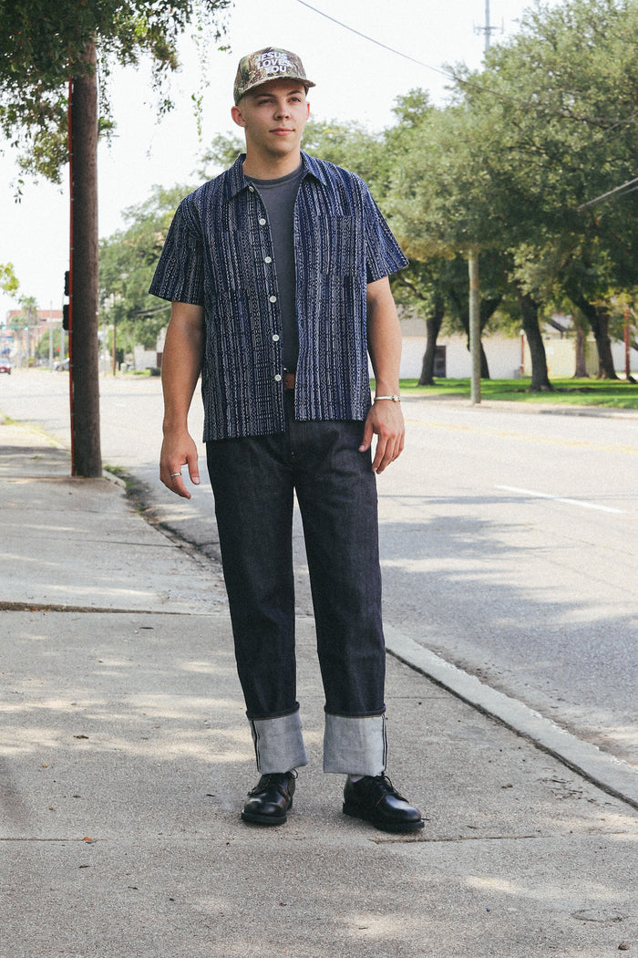 Man wearing dark patterned short-sleeve shirt, cuffed dark jeans, black shoes, and camo cap standing on sidewalk