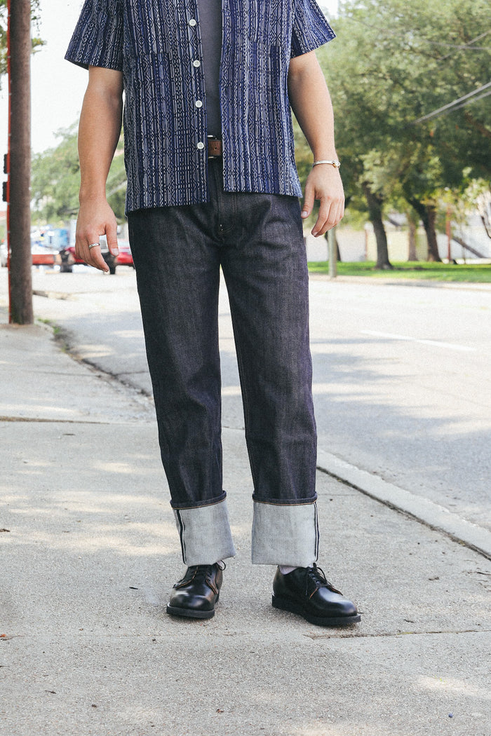 Casual male outfit with cuffed dark denim jeans, black leather shoes, and a patterned short sleeve shirt on city sidewalk