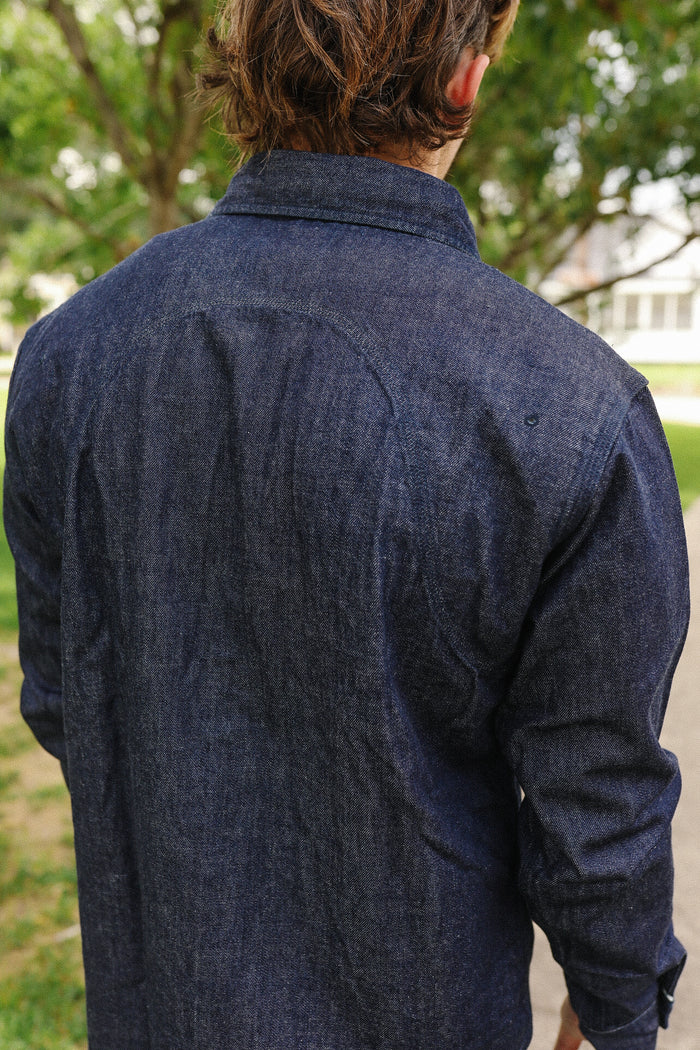 Back view of person wearing indigo denim medical shirt outdoors with greenery and house background