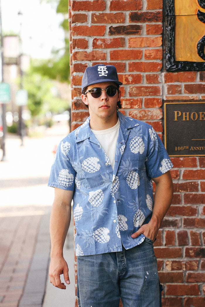 Man wearing Duke Kahanamoku indigo cotton Hawaiian shirt with white pineapple print, sunglasses, and baseball cap leaning against red brick wall