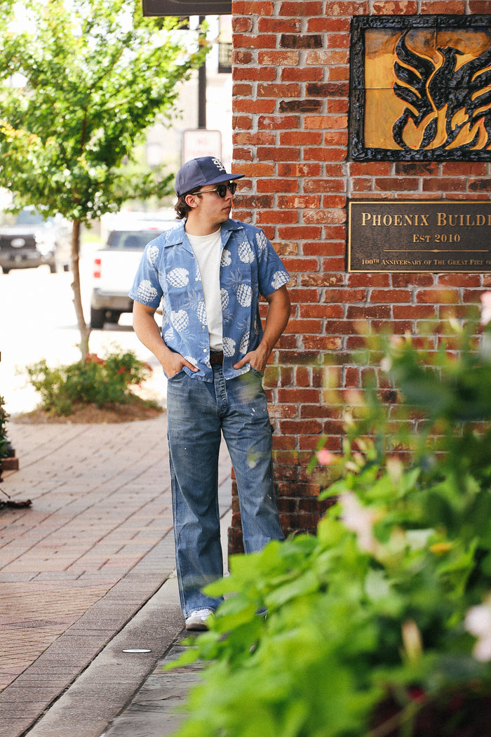 Man wearing indigo blue Hawaiian shirt with white pineapple print, jeans, and cap standing by brick wall plaque