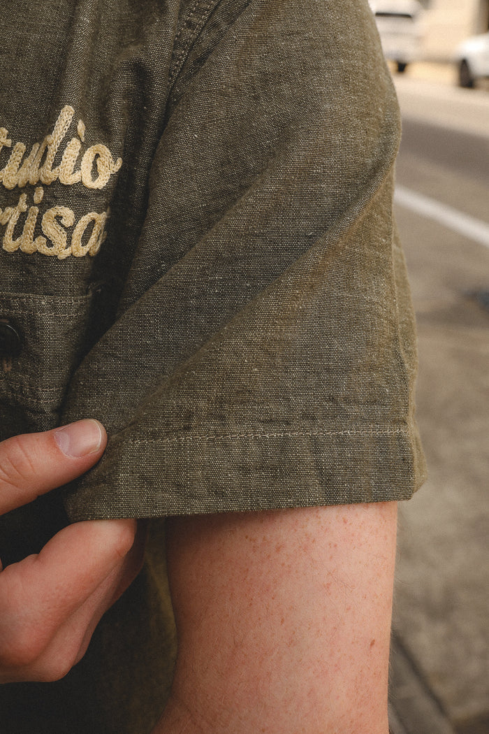 Close-up of a person's hand holding the cuff of an olive green short sleeve work shirt embroidered with 'Studio Artisans'