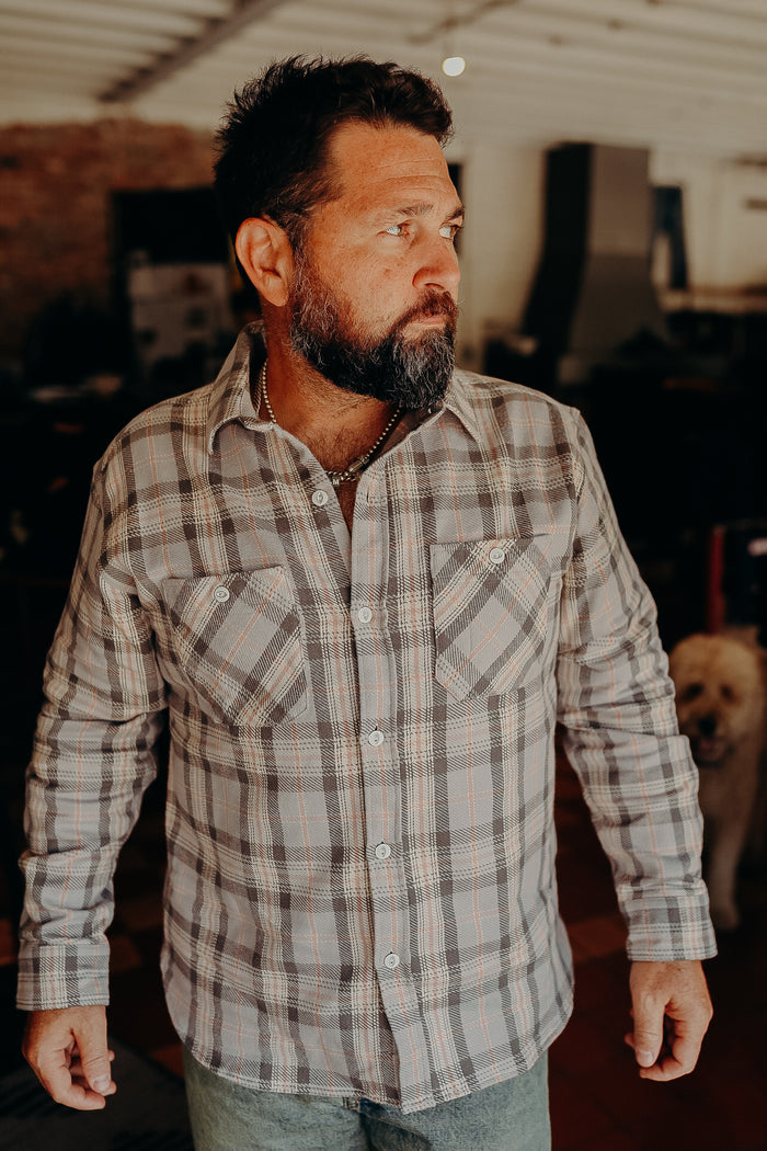 Bearded man wearing a brown and beige plaid button-up shirt indoors with a dog in the background