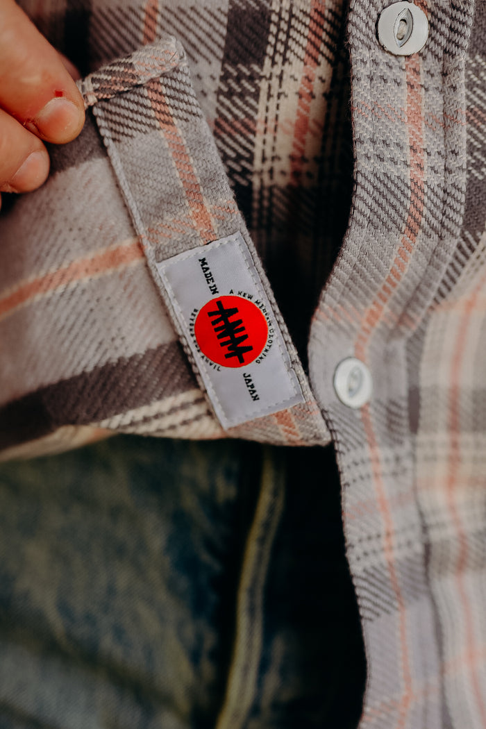 Close-up of hand holding a beige and orange plaid shirt with a Made in Japan label from Iron Shop Provisions