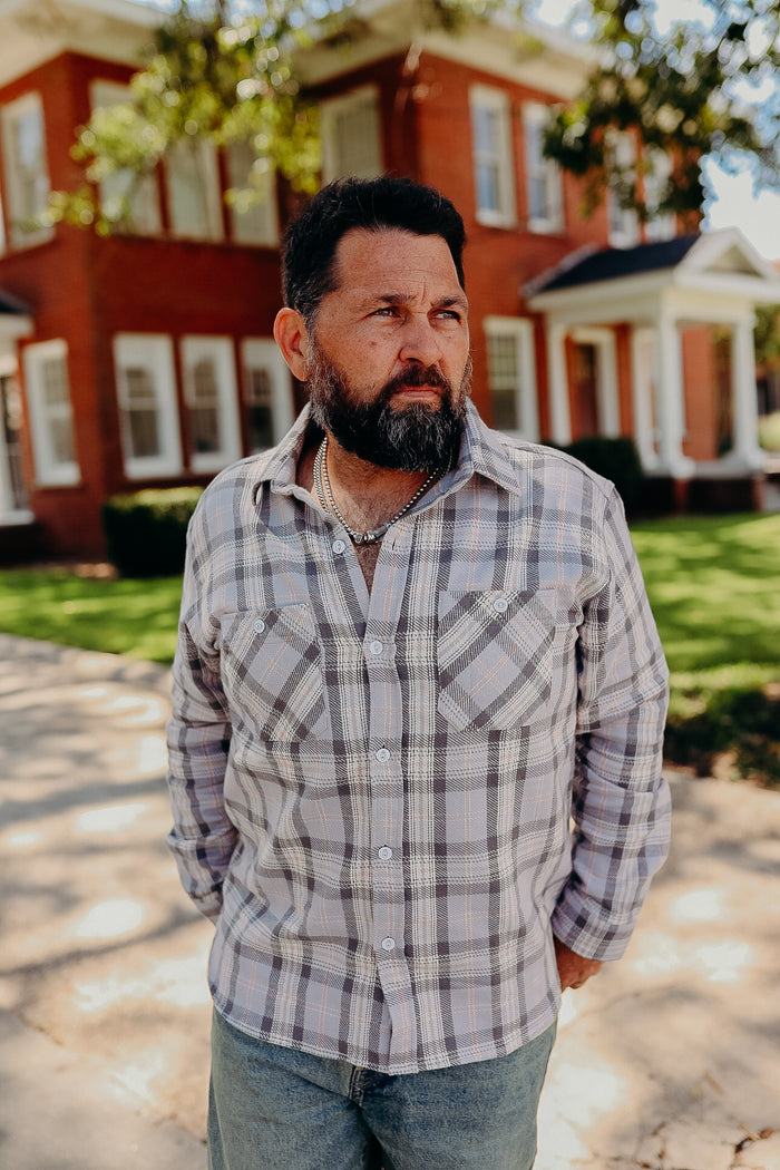Bearded man in plaid button-up shirt and jeans standing outdoors with a brick house background