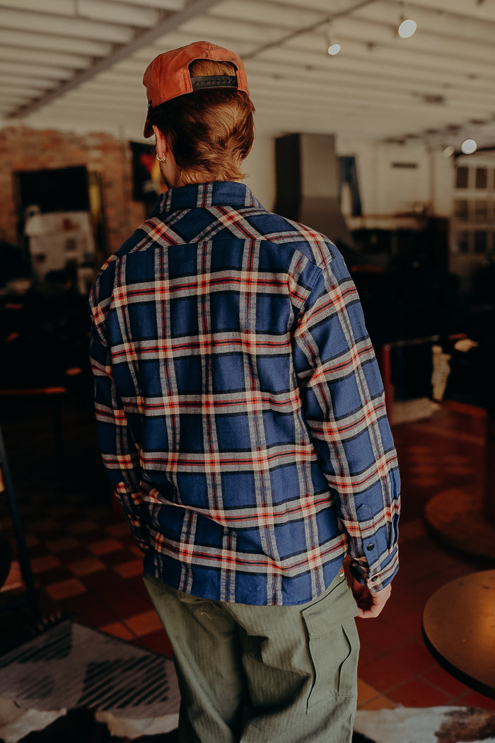 Back view of person wearing blue and red plaid shirt, olive green cargo pants, and orange cap indoors