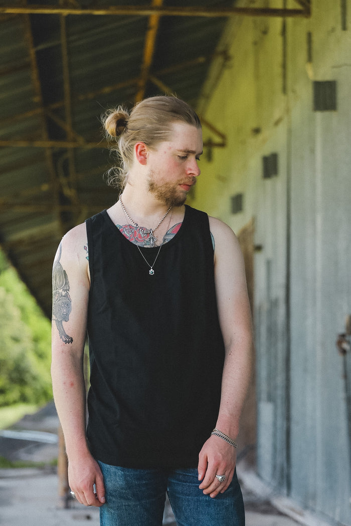 A man with a blonde bun and beard wearing a black sleeveless tank top and blue jeans, standing in an industrial covered walkway.