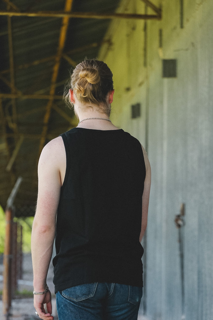 A person viewed from behind wearing a black sleeveless tank top and blue jeans, standing in a covered walkway with a concrete wall on the right.