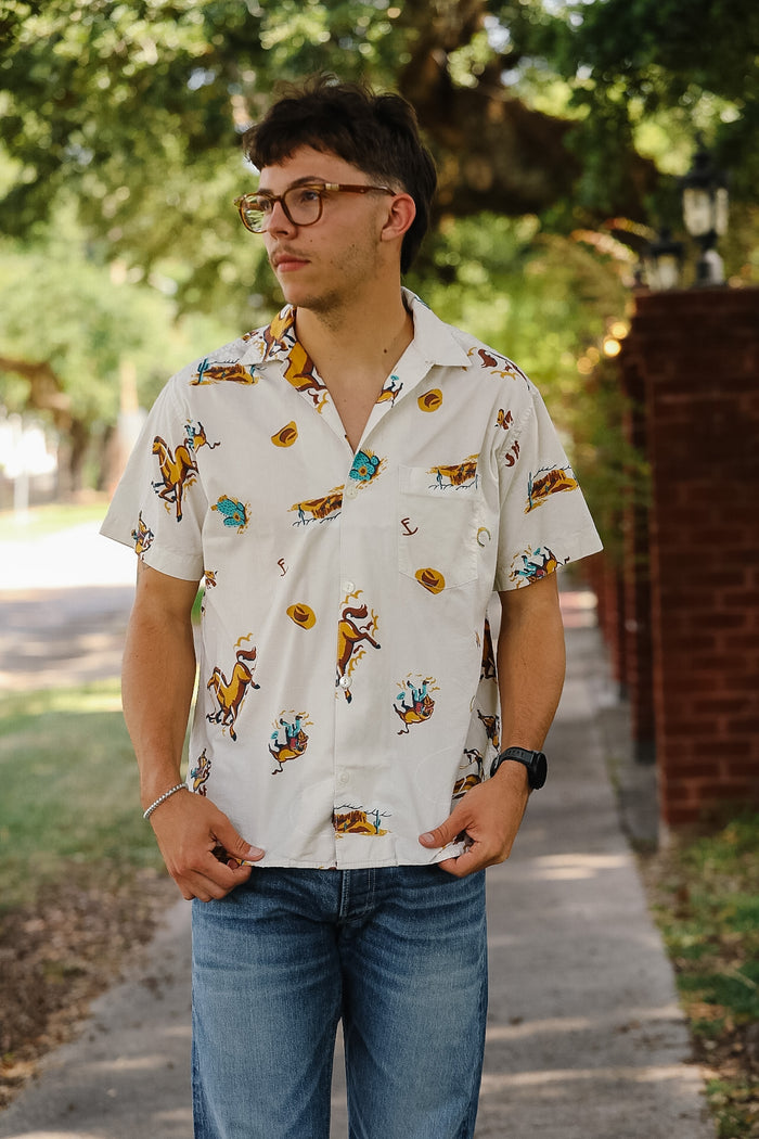 A man wearing a white short-sleeve button-up shirt with a colorful tropical and floral print, paired with blue jeans, standing outdoors on a sidewalk with trees and a brick wall in the background.