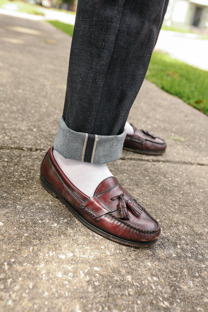 Close-up of rolled-up dark selvedge denim jeans paired with white socks and brown leather tassel loafers on sidewalk
