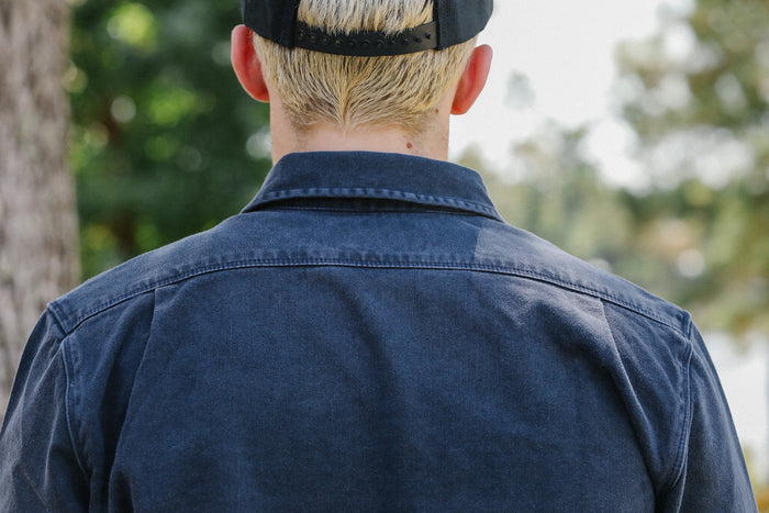 Back view of person wearing dark indigo work shirt and black snapback cap outdoors with blurred trees