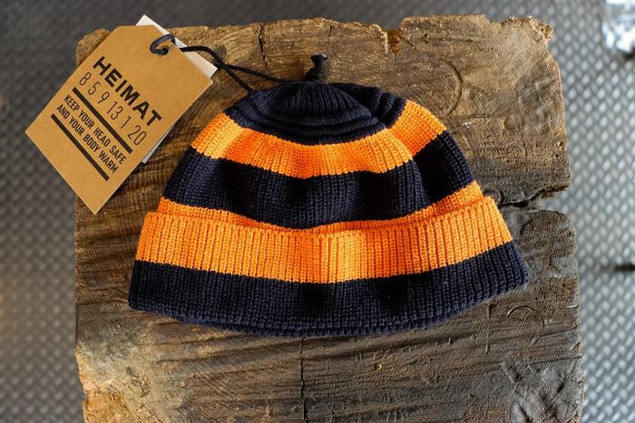 Knitted beanie with orange and black stripes on a wooden block, featuring a 'HEIMAT' tag.
