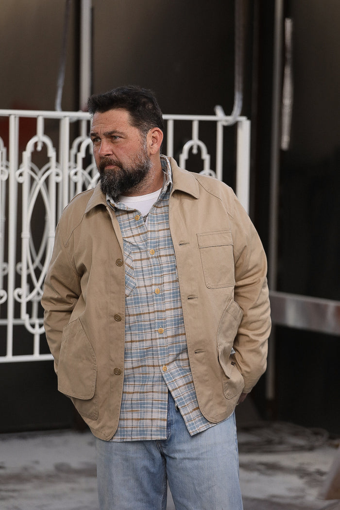 Man with beard wearing beige work jacket and plaid shirt standing outdoors near white iron gate