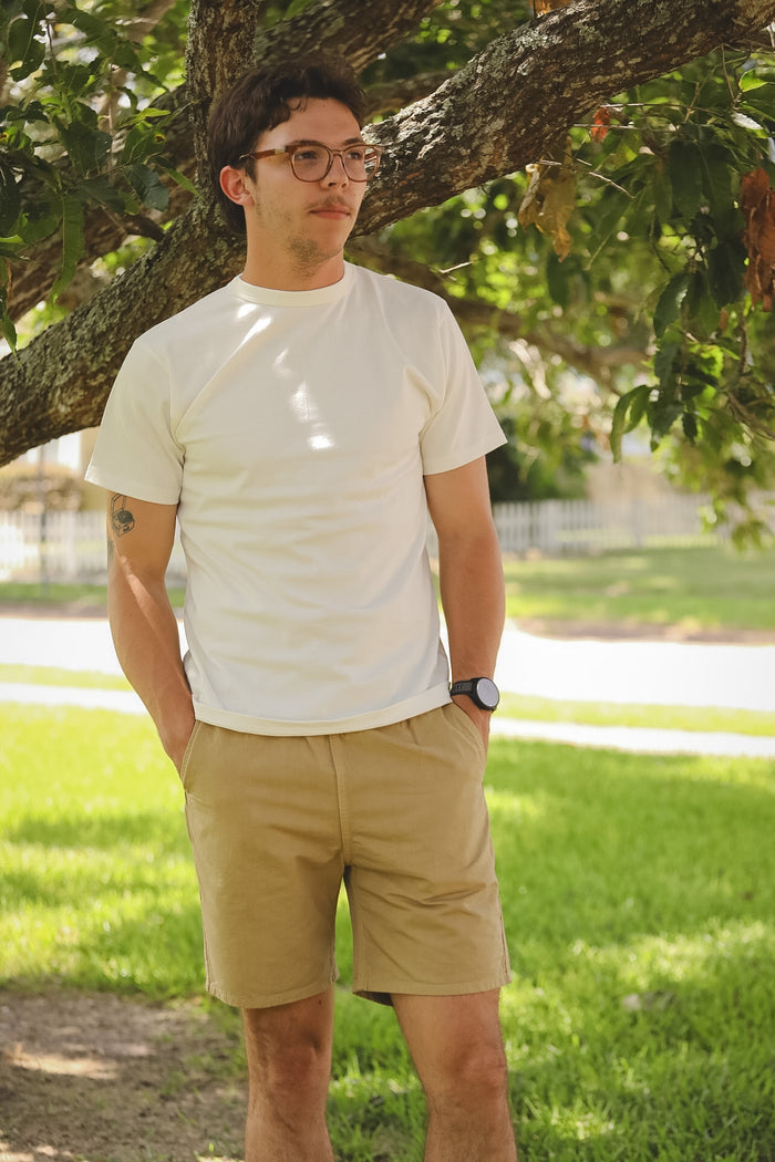 Man wearing a white t-shirt and beige shorts standing under a tree in a park.