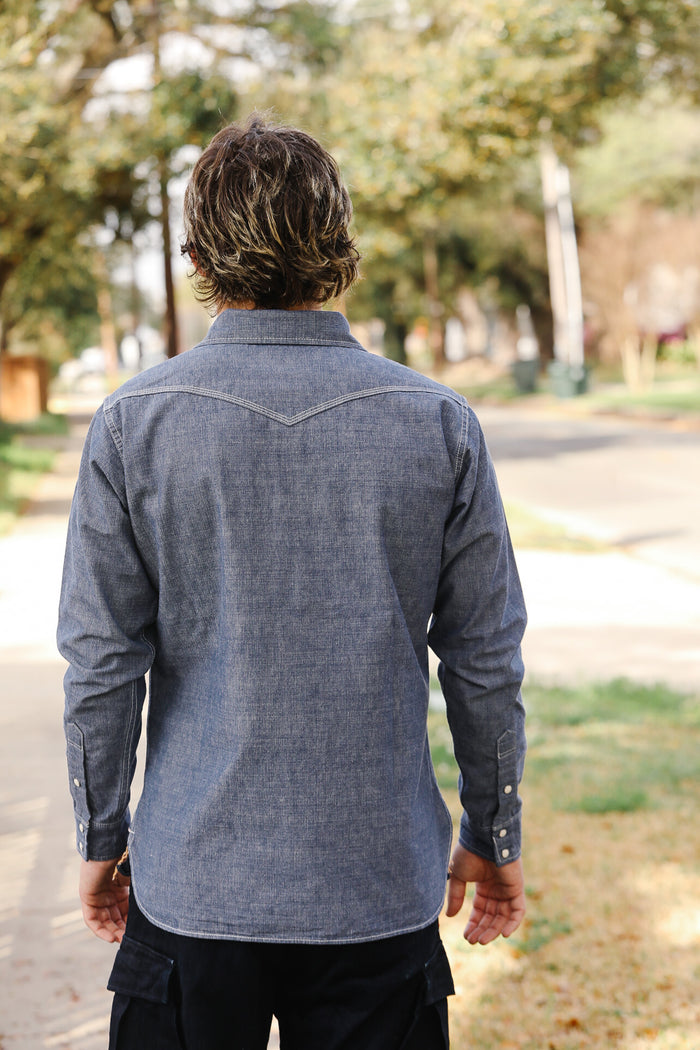 Person wearing a blue denim shirt walking outdoors with trees in the background