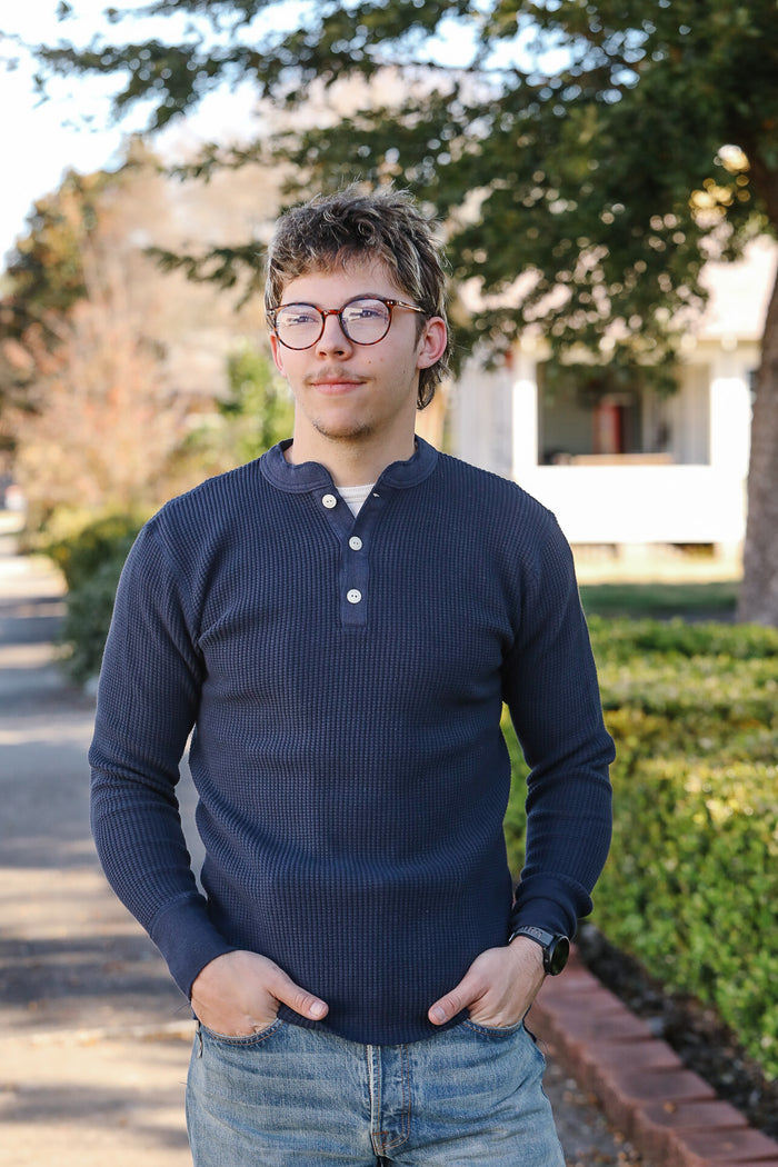 Person wearing a navy blue long-sleeve shirt standing outdoors with trees and a building in the background.