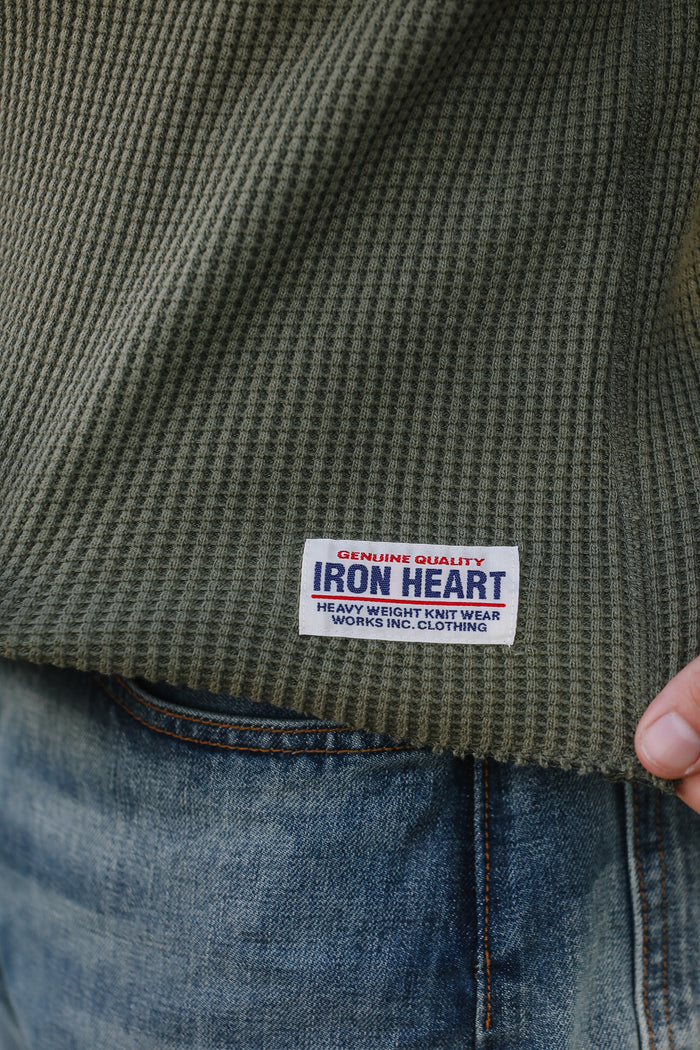 Close-up of a green textured shirt with an Iron Heart label on a person wearing blue jeans.